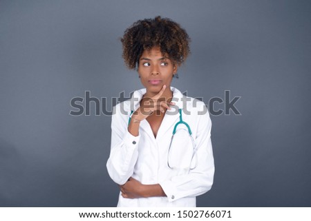 Dreamy African American doctor female with pleasant expression, wearing medical uniform, looks sideways, keeps hand under chin, thinks about something pleasant, poses against gray background.