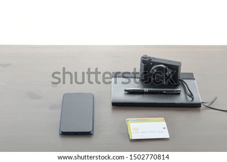 A notebook with a pen on top, payment cards, a mobile phone, a camera on a wooden table with a white background.