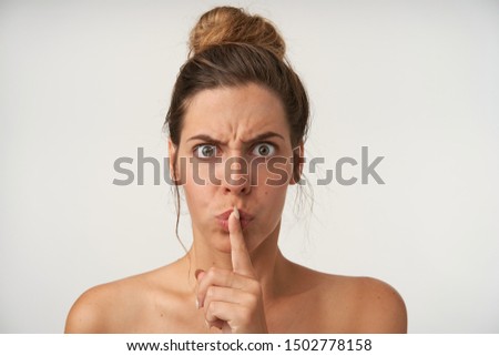Indoor shot of pretty grumpy female raising index finger to her lips, asking to keep silence, frowning and looking seriously to camera