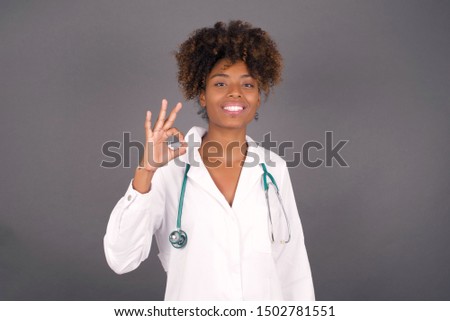Glad attractive African American doctor woman shows ok sign with hand as expresses approval, has cheerful expression being optimistic. Standing against gray wall.