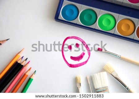 happy smile painted with raspberry paint on a white sheet of paper with colored pencils, a box with paint and brushes for painting. The concept of classes with children. Drawing with children