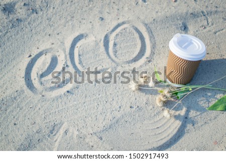 The inscription "eco" on the sand. Paper cup with coffee. Dry flowers on the sand. Bright summer good morning. Brown paper cup with plastic white lid.