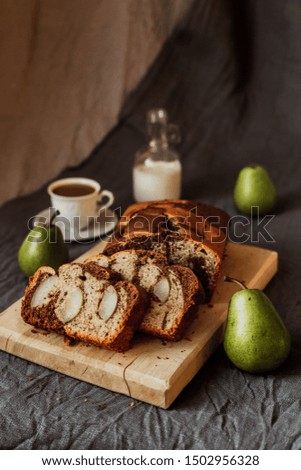 A seasonal cake with pears served on a wooden cutting board, fresh pears, coffee with milk in a white cup, milk in a bottle. Natural light. Vertical.