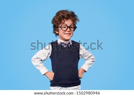 Positive little genius in nerdy glasses and school uniform keeping hands on waist and looking away against blue background