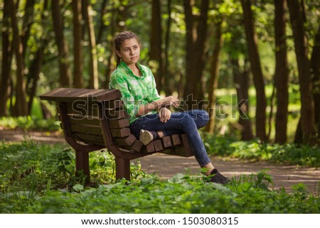 Emotional girl teenager with long hair hairstyle braids in a green shirt sitting on a bench in the park.