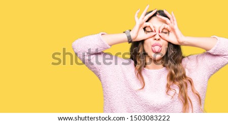Young beautiful woman wearing pink sweater doing ok gesture like binoculars sticking tongue out, eyes looking through fingers. Crazy expression.
