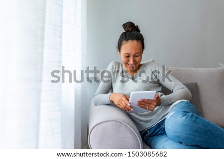 Photo of a mature Asian woman sitting by the window and using digital tablet at home. Pretty woman with digital tablet sitting in sofa at home. Smiling mature woman at home connected on internet