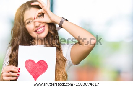 Young adult woman holding card with red heart with happy face smiling doing ok sign with hand on eye looking through fingers