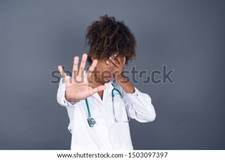 People, body language. Young  African American doctor woman covers eyes with palm and doing stop gesture, tries to hide from everybody. Don't look at me, I don't want to see, feels ashamed or scared.