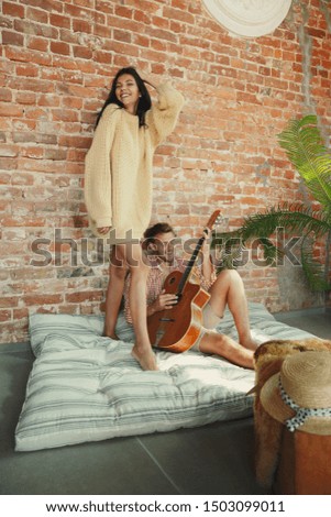Couple of lovers at home relaxing together. Caucasian man playing guitar while woman dancing. Having weekend, looks tender and happy. Concept of relations, family, autumn and winter comfort.