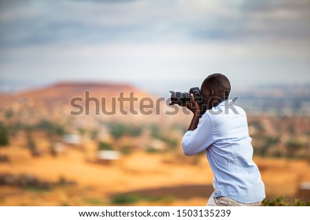 African man taking photos in the Sahel from a higher plateau having a traditional sahelian village in the background at the foothill of flat-top hills outside Niamey capital of Niger