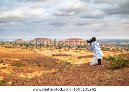 African man taking photos in the Sahel from a higher plateau having a traditional sahelian village in the background at the foothill of flat-top hills outside Niamey capital of Niger