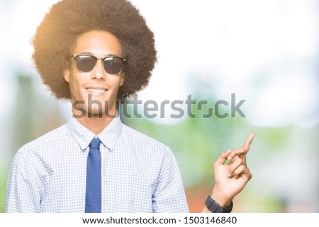 Young african american business man with afro hair wearing sunglasses with a big smile on face, pointing with hand and finger to the side looking at the camera.