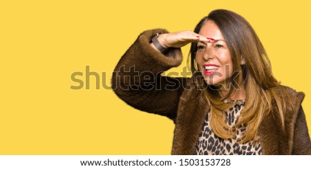 Beautiful middle age elegant woman wearing mink coat very happy and smiling looking far away with hand over head. Searching concept.