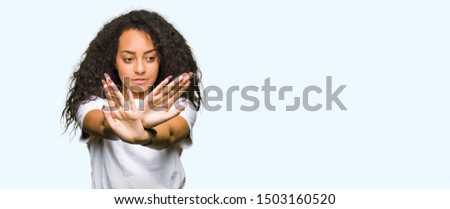 Young beautiful girl with curly hair wearing casual white t-shirt Rejection expression crossing arms and palms doing negative sign, angry face