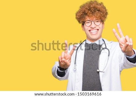 Young handsome doctor man wearing medical coat smiling looking to the camera showing fingers doing victory sign. Number two.