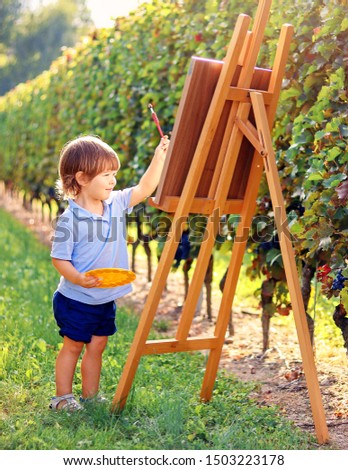 Little toddler boy painting a picture on easel outdoors at nature in sunny vineyards at bright autumn day. Child creativity. Creative hobby and leisure activity. Summer lifestyle.