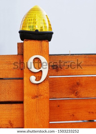 Metallic house number 9, nine and a signal lamp with flashing light on a wooden fence post in front of a white wall.