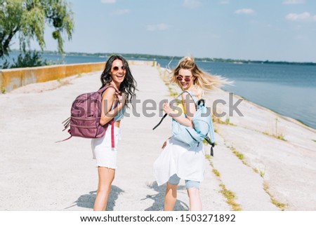 cheerful blonde and brunette girls walking with backpacks near river in summer