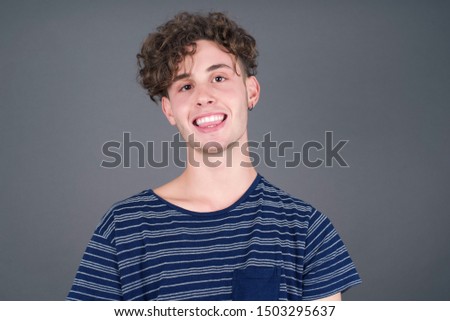 Beautiful Young caucasian man with happy and funny face smiling and showing tongue. Wearing casual clothes and standing against gray studio background.