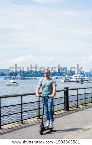 Young American Man traveling in New York City, wearing light green T shirt, blue jeans, sneakers, sunglasses, shoulder carrying back bag, riding on electric scooter, pass by Hudson River with boats.
