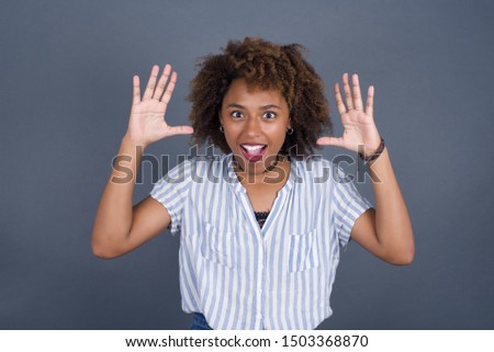 Portrait of beautiful African American female looks with excitement at camera, keeps hands raised over head, notices something unexpected, isolated over gray wall. 
