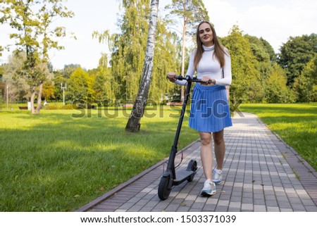 A beautiful girl walks along the path in the park and rolls her electric scooter