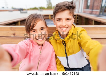 childhood, technology and people concept - happy children or brother and sister sitting on wooden street bench outdoors taking selfie
