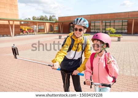 education, childhood and people concept - happy school children in helmets with backpacks and scooters taking picture by smartphone on selfie stick outdoors