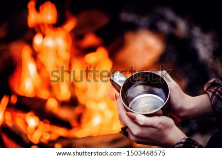 Closeup photo of hands holding camping  cup near bonfire. Ring on fingers. Mug with warm beverage. Drinking water in camp.