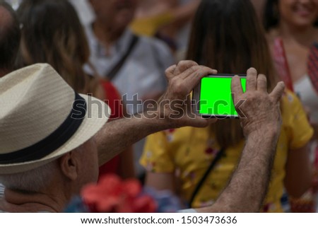 Rear close up of a middle age caucasian man hand taking a picture with a phone held up over a crowd on a blurry background in Malaga in Andalusia in the south of Spain. Green screen on the phone.