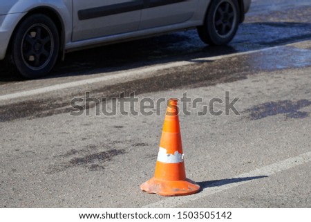 An orange cone stands on the road next to the wheels of a passing car. Life in the city. Daylight