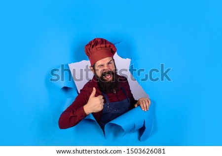 Male chef, cook or baker in hat and apron look through paper hole. Bearded man in chef uniform shows thumb up. Professional happy man chef. Cooking, profession and business. Chef ready to cook dish.