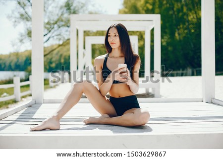 Pretty lady sitting on a summer beach. Girl use the phone; Brunette in a black swimsuit