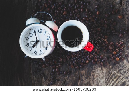 Black coffee in a red ceramic cup with a white alarm clock and coffee beans scattered on the dark wood floor, copy space