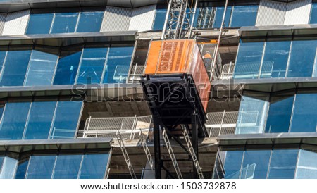 Construction site with elevator. Building with windows and a unrecognisable worker. Useful for building and construction concepts, work health and safety, work force or blue/teal and orange concepts. Royalty-Free Stock Photo #1503732878