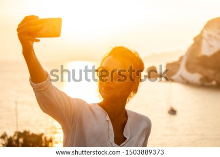Young woman with smartphone taking selfies with sunset in front of the sea on Ponza island coast, on a wall with view of the ocean. 9817