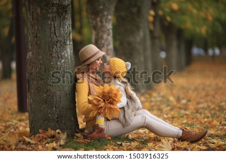 beautiful blonde mother and daughter of European appearance are sitting on the yellow leaves in the park, looking tenderly at each other and kissing, a number of trees looking away