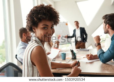 Young afro american woman is looking at camera and smiling while her boss standing near whiteboard and discussing something with team