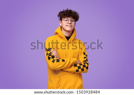 Positive teen boy in yellow hoodie crossing arms and looking at camera against bright violet background Royalty-Free Stock Photo #1503928484