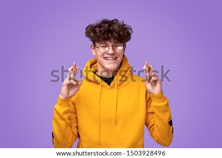 Happy teen boy in nerdy glasses closing eyes and crossing fingers while hoping for luck against purple background