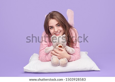Cheerful teen girl in pajama smiling and holding plush bear while lying on soft pillow against violet background