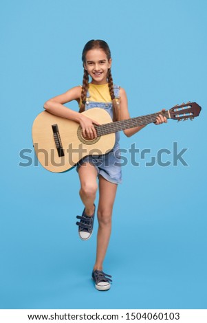 Cheerful little girl in denim overalls playing acoustic guitar and smiling at camera on blue background