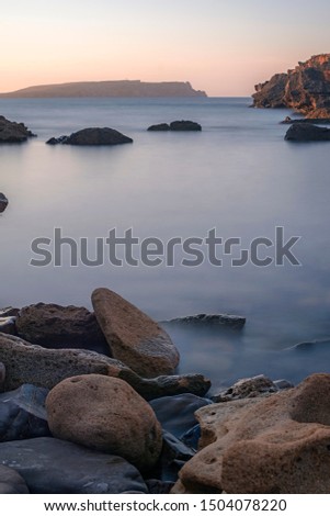 Sunset. Stones in the foreground. Rocks in the sea. Silk effect of long exposure. Mountains on the right and in the background. Blue sea, slightly orange sky.