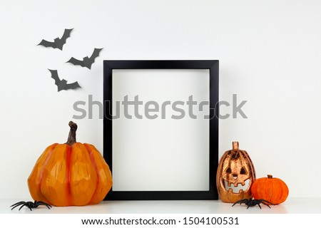 Mock up black frame with Jack o Lantern and pumpkin decor on a shelf or desk. Halloween concept. Portrait frame against a white wall with bats.