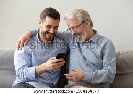 Happy two age generations men family old father embracing young grown adult son having fun enjoying using smart phone bonding watching funny social media video using mobile apps at home sit on sofa Royalty-Free Stock Photo #1504134377