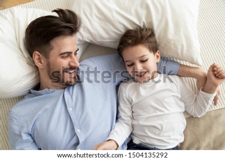 Affectionate family young father embracing cute kid son lying on bed together, little child boy and dad relaxing lay on comfortable mattress in bedroom playing bonding enjoy funny moments, top view