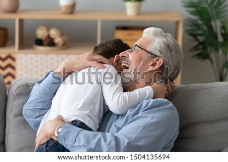 Happy cheerful old senior grandpa embracing cute little boy grandson laughing cuddling at home, two 2 generations family elder grandfather and small grandkid hugging bonding playing sit on sofa Royalty-Free Stock Photo #1504135694