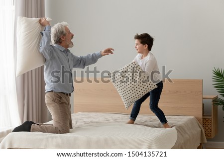 Funny happy two generation men family old grandfather and little cute grandson having fun pillow fight on bed, senior grandpa playing funny game enjoy leisure activity with grandchild boy in bedroom