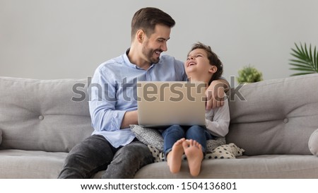 Happy father and little cute son laughing enjoying using laptop together relaxing on family leisure technology lifestyle sit on sofa, smiling dad embracing kid boy having fun with computer at home